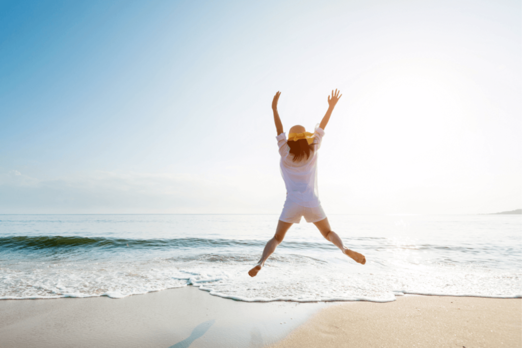 woman jumping on beach with arms in the air, looking hapy